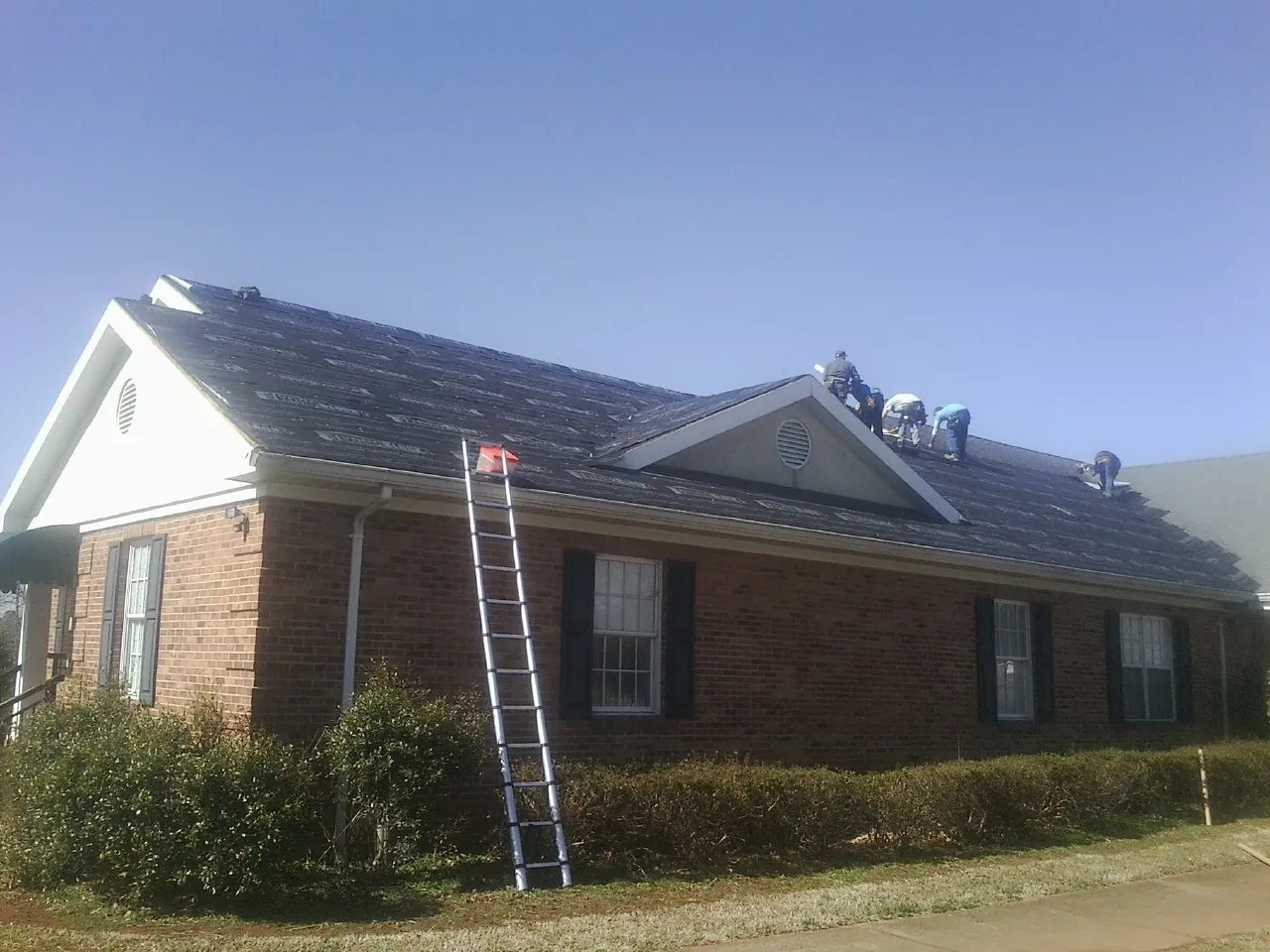 Crew tearing shingles off a brick ranch roof with a ladder set — Emergency Roof Repair work by our crew in North Webster