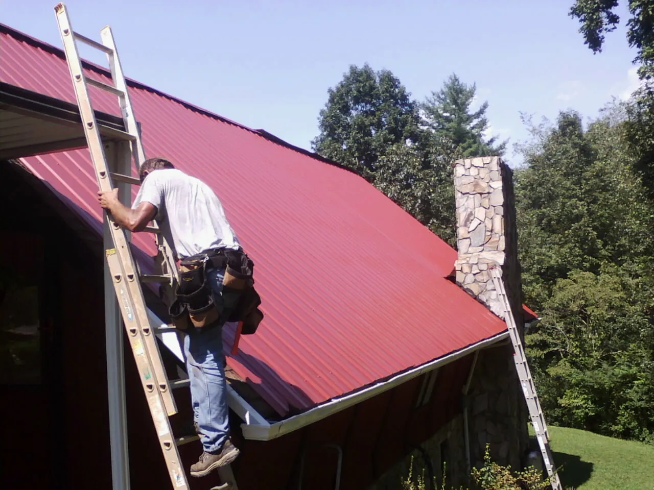 Roofer on a ladder beside a red metal roof and stone chimney — Soffit & Fascia Repair by Roofing Brothers in North Webster